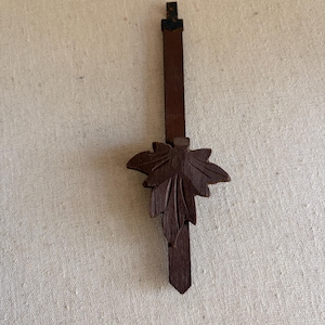 May include: A dark brown wooden clock hand featuring a carved maple leaf ornament attached to the center. The vertical component is a long, narrow strip with a pointed bottom tip and a small, dark metal connector at the top end of the piece.