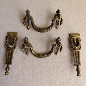 May include: An assortment of antique brass furniture hardware, including two curved handles and two vertical decorative pieces. The hardware features ornate detailing and a warm, aged patina, suggesting a vintage aesthetic. The pieces are arranged on a neutral background.