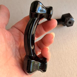 May include: Two black, curved cabinet pulls with a glossy finish. Each pull has a rounded handle and decorative ends. One pull is held in a hand, while the other is in the background. The pulls are likely for furniture.