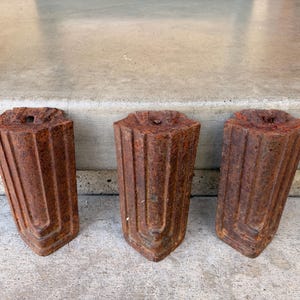 May include: Three weathered, rust-colored metal architectural elements. Each piece has a rectangular shape with a decorative, fluted design and a hole at the top. The objects are arranged in a row, showcasing their aged appearance and unique form.