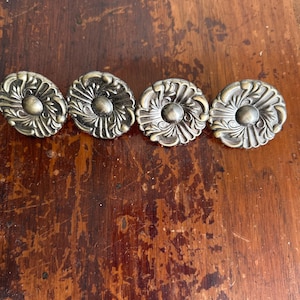 May include: Four antique bronze-colored cabinet knobs with a floral design. Each knob has a central rounded element surrounded by detailed, stylized leaves. The knobs are arranged in a row on a distressed wooden surface.