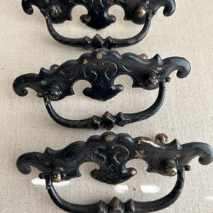 May include: Three antique black drawer pulls with ornate, curved designs. The pulls have a decorative, scrolled top and a curved handle. The metal has a distressed finish with hints of gold, suggesting age and wear. These vintage pulls are ideal for furniture restoration.
