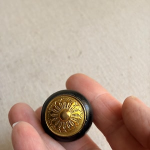 May include: A round cabinet knob featuring a smooth black outer ring and a central gold-colored metal inlay. The gold inlay displays a detailed, embossed radial pattern with a small, raised circular dome at its very center.