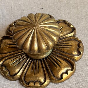 May include: A brass drawer knob featuring a circular, fluted top piece attached to a flat, flower-shaped backplate. The metal has a warm, aged gold tone with dark recessed areas between the raised petals and ridges, creating a textured appearance.