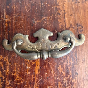 May include: Antique brass drawer pull with a decorative design. The pull features a curved handle and ornate backplate with a central flourish. The brass has a weathered appearance, suggesting age and use. Ideal for vintage furniture restoration projects.