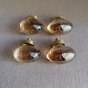May include: Four oval-shaped cabinet knobs with a clear, amber-colored glass body and a gold-colored metal base. The knobs are arranged in a square pattern against a neutral-colored fabric background. These knobs are suitable for furniture or cabinet upgrades.