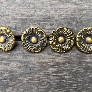May include: Four antique brass cabinet knobs with a floral design. Each knob features a central rounded element surrounded by detailed, swirling patterns. The knobs are arranged in a row against a weathered wooden surface.
