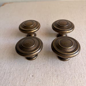 May include: Four antique bronze cabinet knobs with a circular, ridged design. The knobs have a warm, aged appearance, suggesting vintage or retro styling. These knobs are suitable for furniture restoration or adding a classic touch to cabinetry.