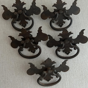 May include: Five antique bronze drawer pulls with a decorative leaf and scroll design. Each pull features a circular handle and ornate backplate. These vintage hardware pieces are perfect for adding a touch of rustic charm to furniture.