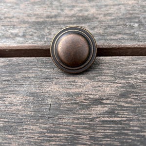 May include: A circular metal button with a dark bronze finish. The button features a smooth, raised center dome surrounded by a recessed ring and a slightly elevated outer rim, creating a layered, concentric design with a weathered, metallic texture.