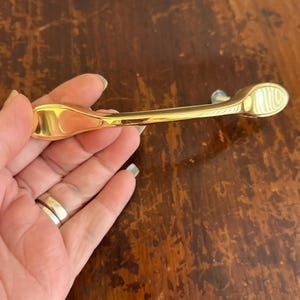 May include: A gold-colored cabinet pull with a curved design. The pull has a smooth, polished finish and is held in a hand. The cabinet pull is likely for a drawer or cabinet door.