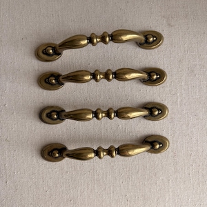 May include: Four antique brass drawer pulls. Each pull has a curved handle with decorative detailing and circular end plates. The pulls are arranged horizontally against a neutral background, showcasing their aged, golden finish.