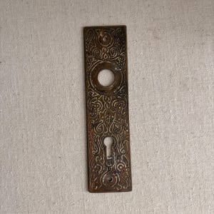 May include: Ornate antique door plate with a keyhole and a circular opening. The rectangular plate is made of a bronze-colored metal and features an intricate floral design. The plate is likely from the early 20th century.