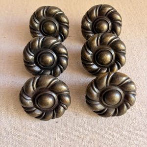 May include: Six antique bronze-colored cabinet knobs with a floral design. Each knob has a central circular element surrounded by a series of curved ridges, creating a flower-like appearance. The knobs are arranged in two rows of three.
