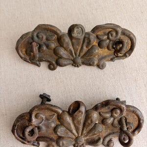 May include: Two antique bronze drawer pulls with a floral and scroll design. Each pull features a central fan-shaped element and decorative swirls. The pulls are attached with screws and show signs of age and wear.