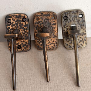 May include: Three metal wall hooks with rectangular backplates featuring a hammered, textured finish. Each hook has a central vertical arm extending outward. The finishes vary in tone from dark bronze to a lighter, muted brass color.