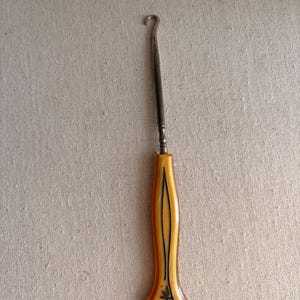 May include: An antique crochet hook with a curved metal end and a handle. The handle is a light amber color with black accents and a decorative floral design at the base. The hook is a vintage craft tool.