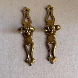 May include: Two matching vertical brass drawer pulls with a polished metallic finish. Each pull features an elongated, symmetrical shape with open cutouts at the top and bottom, and a central horizontal bar handle attached to a decorative plate.