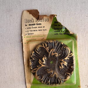 May include: A vintage package of drawer knob back plates. The back plate is a dark bronze color with gold accents and a floral design. The package reads "Back Plates" and "California Styled".
