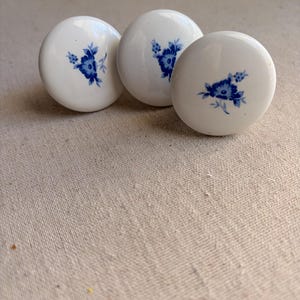 May include: Three round white ceramic knobs, each featuring a small blue floral pattern painted in the center. The knobs have a smooth, glossy finish and a domed shape, designed for use as hardware on furniture such as drawers or cabinets.