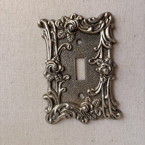 May include: A rectangular silver-toned metal light switch plate featuring a raised relief design of roses and scrolling vines. The central area has a textured, stippled finish with a rectangular cutout for a toggle switch and two screw holes.