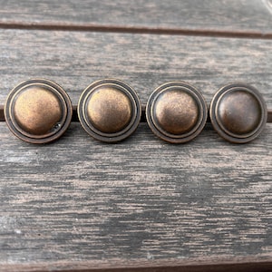 May include: Four antique bronze-colored cabinet knobs with a circular design. Each knob has a raised center and a decorative outer ring. The knobs are arranged in a row on a weathered wooden surface.