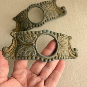 May include: Two antique bronze-colored metal decorative hardware pieces. Each piece has a central circular opening, ornate scrollwork, and a beaded border. The pieces have a weathered, aged appearance, suggesting they are vintage or reclaimed.