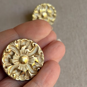 May include: Two ornate, round, antique-style cabinet knobs. The knobs are painted in a cream color with gold accents, featuring a detailed floral design. One knob is in focus, held in a hand, with another slightly blurred in the background.