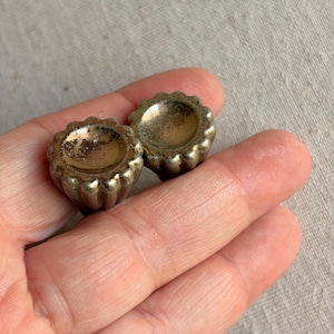 May include: Two small, round metal knobs with a scalloped, fluted edge design. The surface of the knobs has a tarnished, aged silver-tone finish with visible brown oxidation and wear, featuring a concave center and a ribbed, petal-like exterior.