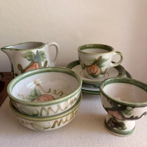 May include: A ceramic set featuring a creamer pitcher, two stacked bowls, a cup with a saucer, and a pedestal bowl. Each piece is off-white with hand-painted green rims and orange fruit motifs with green leaves, showcasing a rustic, glazed finish.