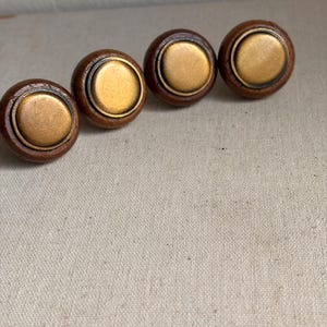 May include: Four circular cabinet knobs arranged in a row. Each knob features a dark brown wooden outer ring surrounding a smooth, matte gold-toned metal center disc. The design is consistent across all four pieces, showcasing a combination of natural wood grain and metallic finish in a simple, rounded shape.
