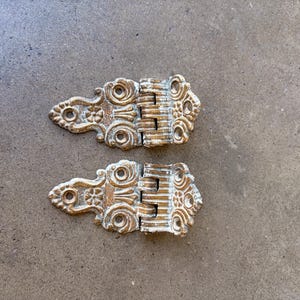 May include: Two antique-style hinges with ornate detailing. The hinges are made of metal with a gold finish and white accents. Each hinge has a decorative floral design on the ends and a series of vertical bars in the center.