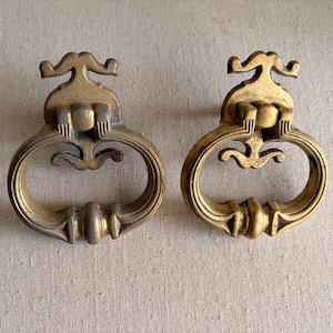 May include: Two antique brass drawer pulls. Each pull features an oval handle with decorative detailing and a stylized top. The brass has a warm, aged patina, suggesting vintage or retro design. The pulls are shown against a neutral background.