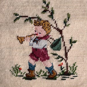 May include: A cross-stitch depiction of a young person playing a horn, standing near a tree with a green bag. The figure wears a white shirt, burgundy shorts, blue socks, and brown boots. The background is a beige fabric.