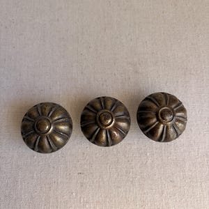 May include: Three antique brass cabinet knobs with a floral design. Each knob has a central circular element surrounded by a series of curved ridges. The knobs are arranged in a horizontal line against a neutral background.