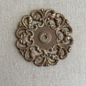 May include: Ornate, circular metal furniture hardware with a central hole. The piece features a brown base with gold-colored accents and a decorative, scroll-like design. The hardware is likely for a cabinet or drawer.
