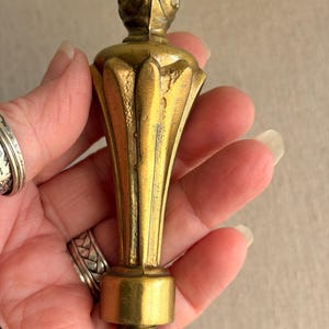 May include: A vintage brass bottle stopper with a decorative, fluted design. The stopper has a rounded top and a cylindrical base. The brass has a warm, aged patina, suggesting it is an antique or vintage item. The stopper is held in a hand.