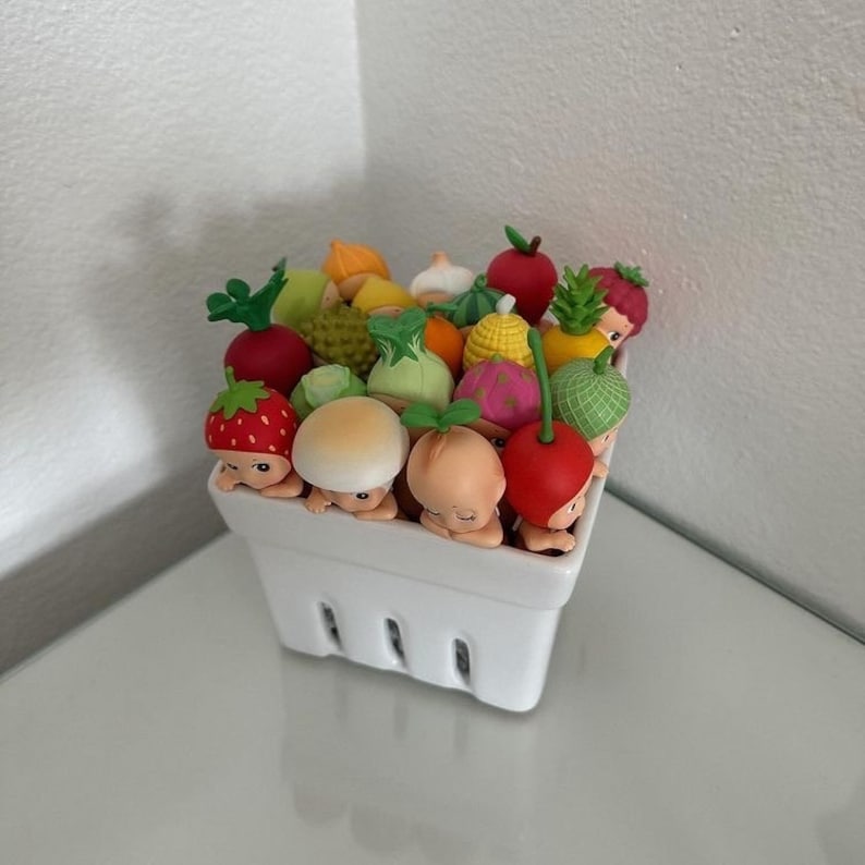 May include: A white ceramic basket filled with miniature fruit and vegetable figurines. The figurines include strawberries, mushrooms, peaches, and cherries, each with a small face peeking over the edge. Other fruits and vegetables are also visible.