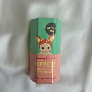 May include: A hexagonal box with a green top and a pink bottom. The box features a cartoon illustration of a baby with deer antlers. Text on the box reads "Sonny Angel HIPPERS. Decorative Mini Figures" and "Attach Me."