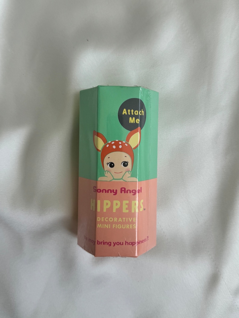 May include: A hexagonal box with a green top and a pink bottom. The box features a cartoon illustration of a baby with deer antlers. Text on the box reads "Sonny Angel HIPPERS. Decorative Mini Figures" and "Attach Me."
