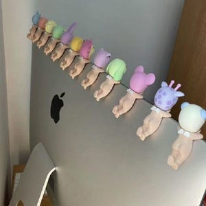May include: A line of small, quirky figurines, each showing a baby-like figure with an animal-themed head. The figurines are in pastel shades and are attached to a silver computer monitor.