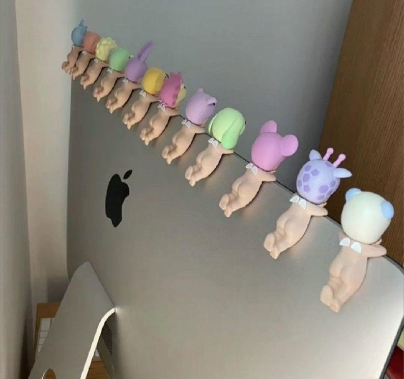May include: A line of small, quirky figurines, each showing a baby-like figure with an animal-themed head. The figurines are in pastel shades and are attached to a silver computer monitor.