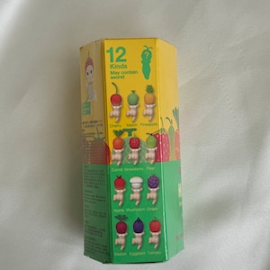 May include: A yellow hexagonal box with illustrations of small figures with fruit and vegetable heads. The box reads "12 Kinds May contain secret" and lists the fruits and vegetables: cherry, melon, pineapple, carrot, strawberry, pear, apple, mushroom, grape, radish, eggplant, and tomato.