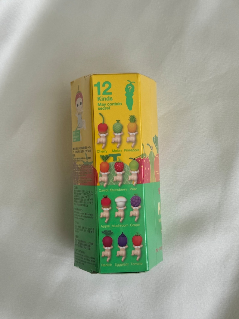 May include: A yellow hexagonal box with illustrations of small figures with fruit and vegetable heads. The box reads "12 Kinds May contain secret" and lists the fruits and vegetables: cherry, melon, pineapple, carrot, strawberry, pear, apple, mushroom, grape, radish, eggplant, and tomato.