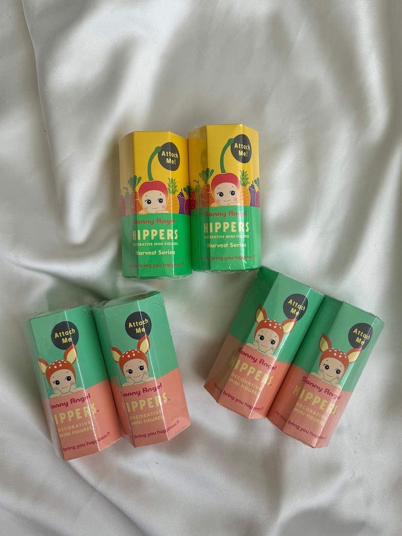 May include: A collection of Sonny Angel Harvest Series decorative mini figures are shown. The packaging is in shades of yellow, green, and peach, featuring a baby with a vegetable-themed hat. The text "Attach Me!" is visible on the boxes.