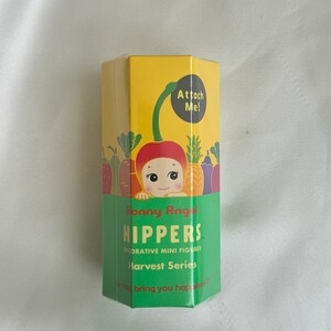 May include: A yellow and green box of Sonny Angel Hippers Harvest Series decorative mini figures. The box features a cartoon illustration of a cherry with a face and the text "Attach Me!". The box also has the text "He may bring you happiness!"
