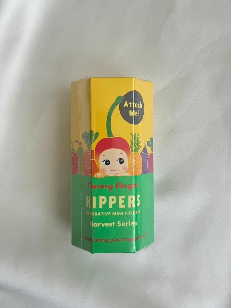 May include: A yellow and green box of Sonny Angel Hippers Harvest Series decorative mini figures. The box features a cartoon illustration of a cherry with a face and the text "Attach Me!". The box also has the text "He may bring you happiness!"