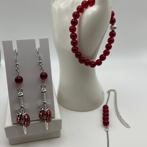 May include: A set of gothic-themed jewelry featuring red beaded bracelets, earrings, and a bookmark. The earrings and bookmark have a sword and heart design. The bracelet is displayed on a white hand-shaped display.