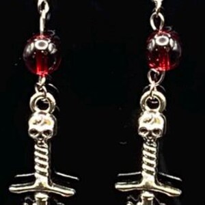 May include: A pair of silver-toned earrings with a gothic theme. Each earring features a red bead, a skull atop a sword, and a red heart pierced by the sword. The earrings hang from silver hooks.