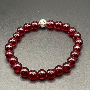 Könnte beinhalten: Ein Armband mit tiefroten, runden Perlen. Es hat auch eine silberfarbene, dekorative Perle. Die Perlen sind zu einem Kreis aufgereiht. Das Armband wird auf einer dunklen Oberfläche präsentiert.